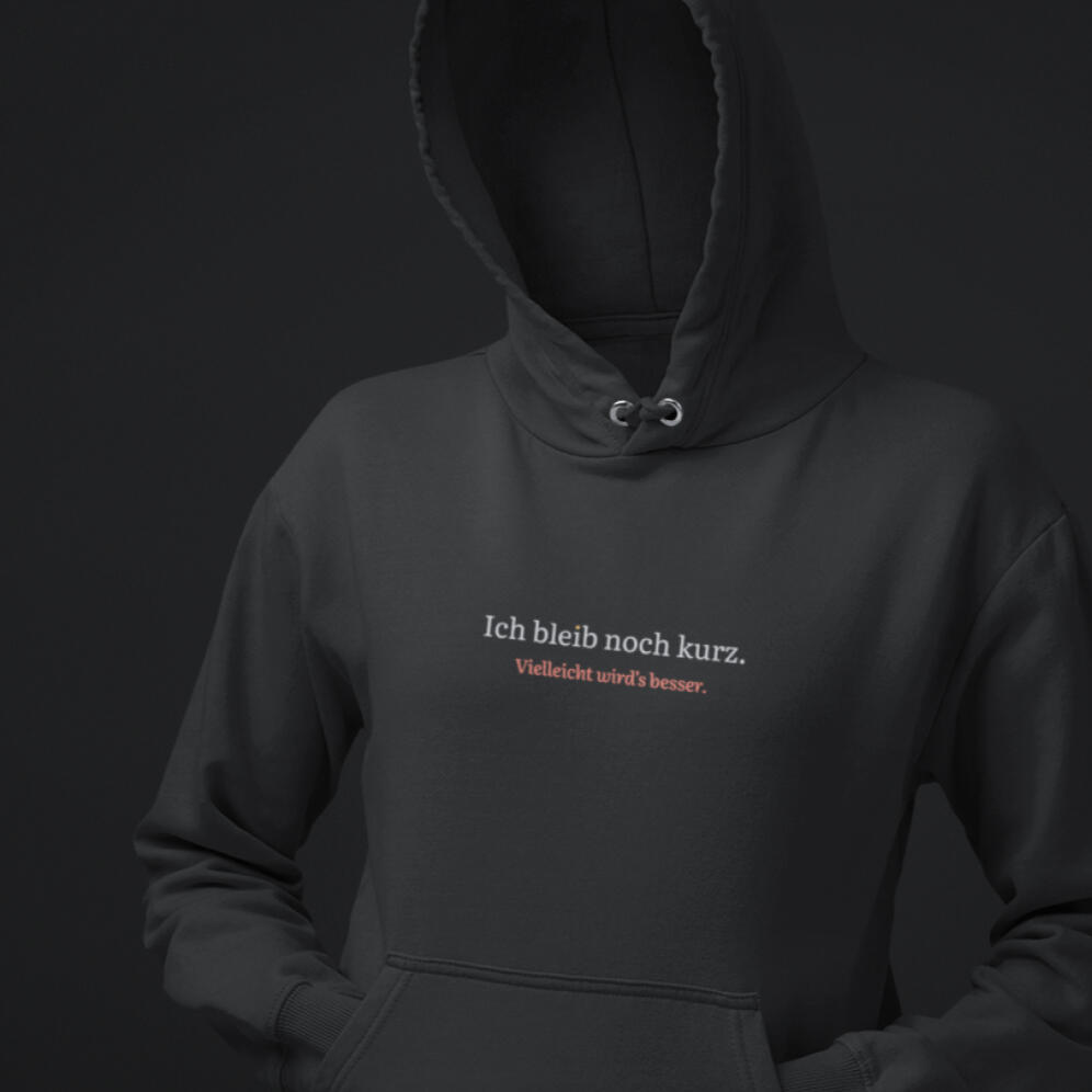 Schwarzer Hoodie mit dem Aufdruck „Ich bleib noch kurz. Vielleicht wird’s besser.“ in weißer und roter Schrift.