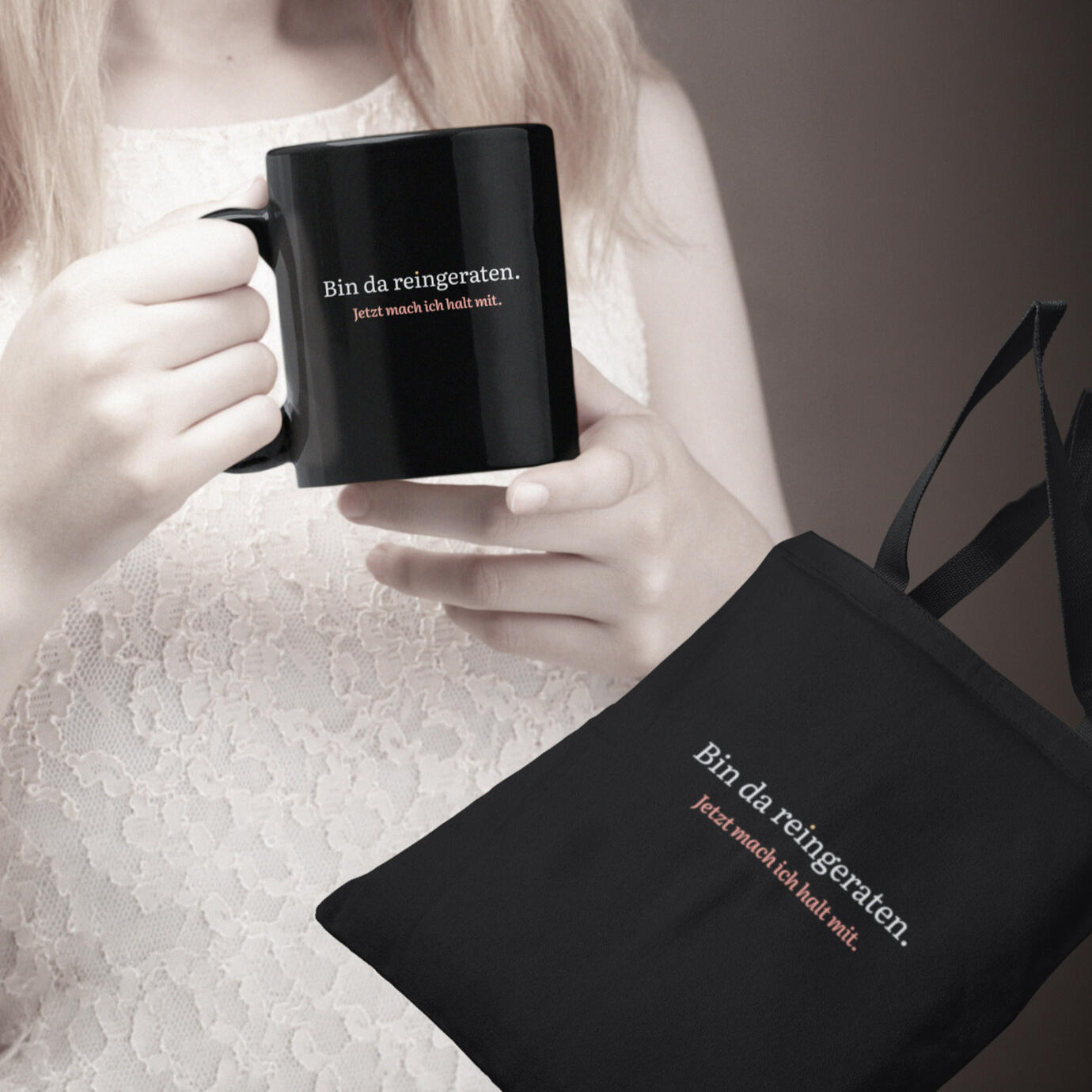 Fotoszene: Person hält Tasse, daneben schwarzer Stoffbeutel, beide mit dem Aufdruck „Bin da reingeraten. Jetzt mach ich halt mit.“